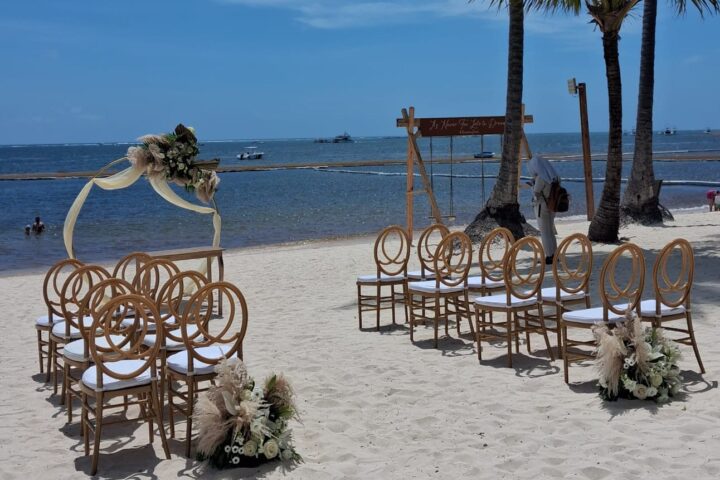 A wooden arch trimmed with dried pampas stands before rattan chairs on bright sand.