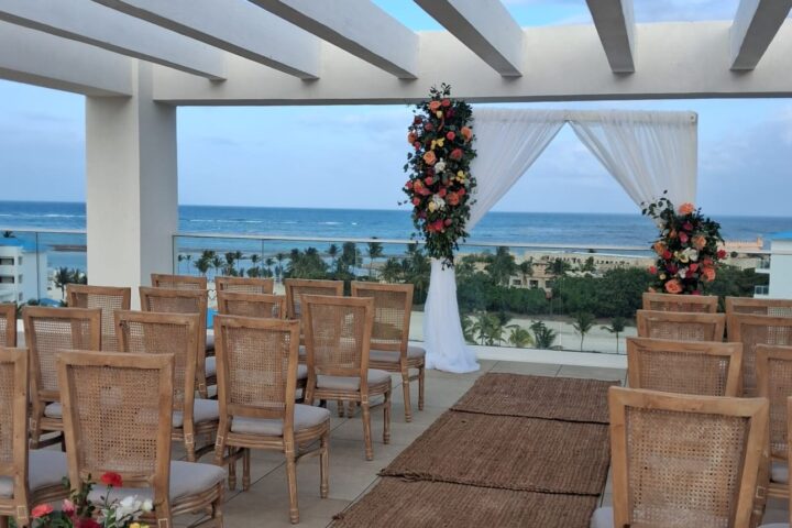 Chairs and a draped floral arch fill a rooftop terrace overlooking the ocean at dusk.